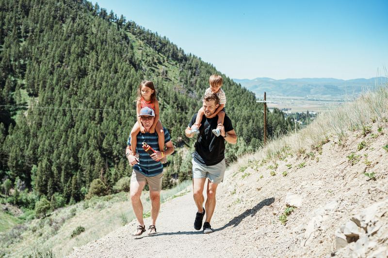 Two dads walking a mountain trail with their kids on their shoulders